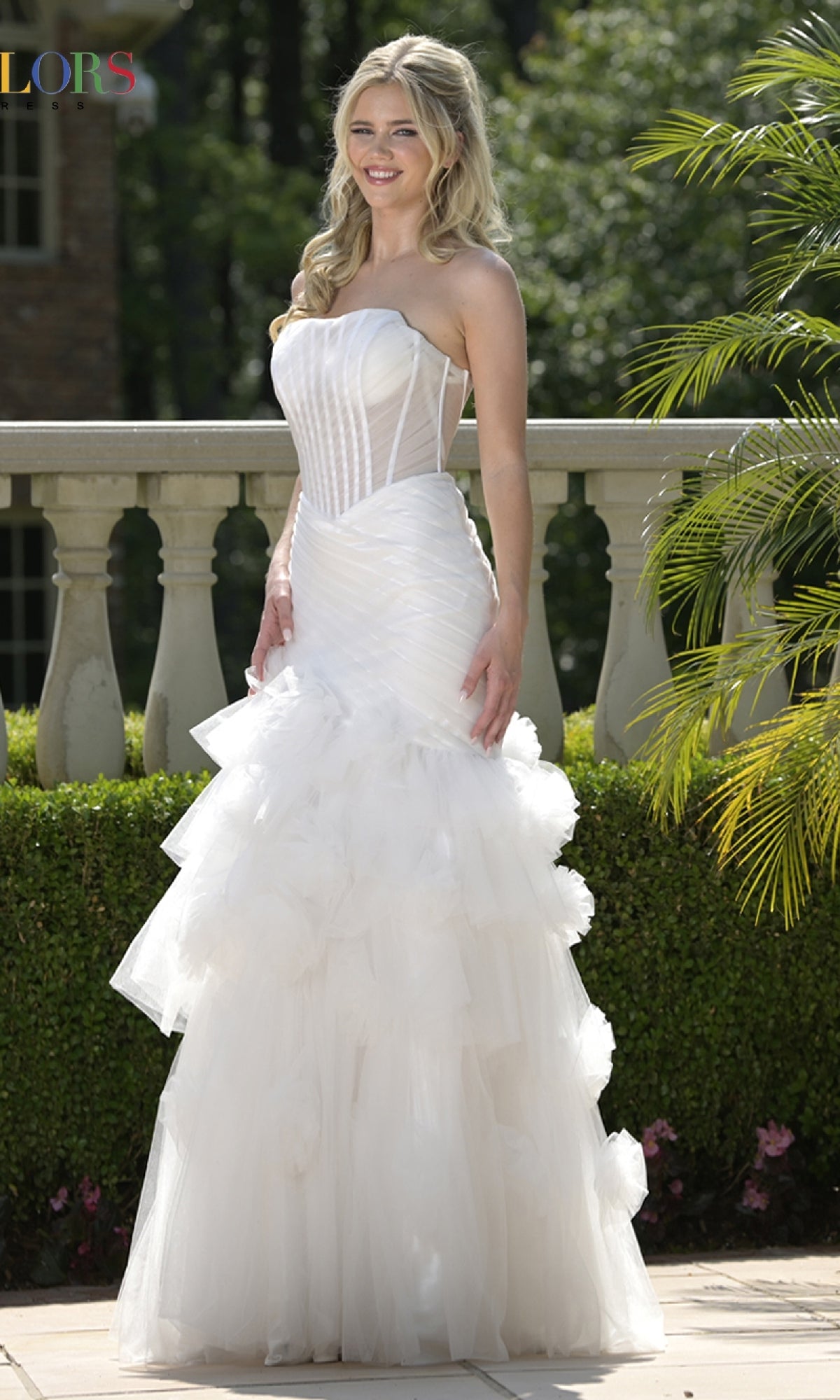 A woman stands outdoors near a stone railing in the Colors Dress 3800 long prom dress, featuring a strapless fitted bodice, lace-up open back, and tiered tulle skirt, smiling amid lush greenery.